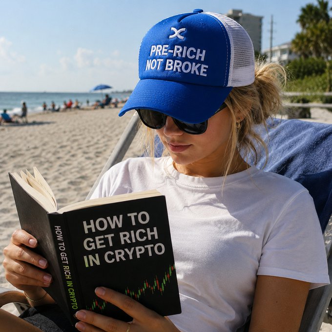 A woman relaxes on a beach reading "How to Get Rich in Crypto" while wearing a blue "Pre-Rich Not Broke" trucker hat, humorously visualizing the grind toward future wealth that the caption "keep grinding, you'll get there" encourages.