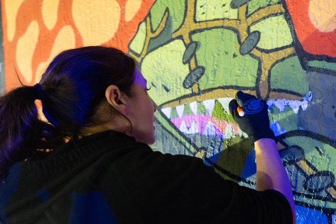 A woman with dark hair tied back paints a vibrant, colorful graffiti mural on a wall, wearing black gloves while working on a green and yellow creature design.