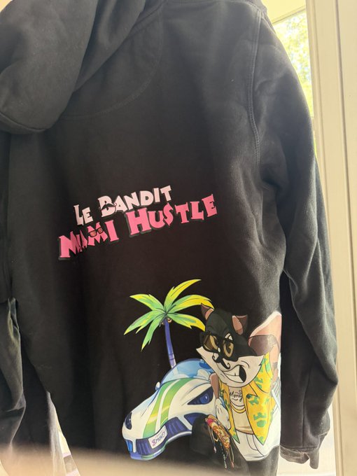 A black hoodie features the text "LE BANDIT" and "HUSTLE" above a colorful cartoon graphic of a cat bandit character wearing a Hawaiian shirt, gold chain, and cap, posed next to a sports car and palm tree. The image relates to the post asking Hacksaw Gaming for an exclusive slot featuring "Le Bandit," matching the hoodie's theme of the popular mascot character.