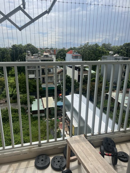 A view from a balcony overlooking a residential neighborhood with houses, trees, and rooftops, featuring a wooden bench and weight plates on the tiled floor in the foreground.
