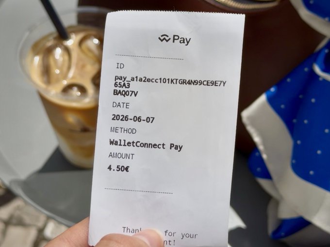 The image shows a person holding a receipt that has been used to pay for their coffee at a cafe. The receipt displays the details of the payment made, including the amount spent and the date of purchase. There are also some text elements on the receipt, such as "Paid" and "@ WalletConnect Pay". This suggests that the person paid using a mobile wallet or an online payment system to cover their coffee expenses at the cafe. The image also includes a photo of a cup of iced coffee with ice cubes in it, which adds context to the scene and highlights the payment process.