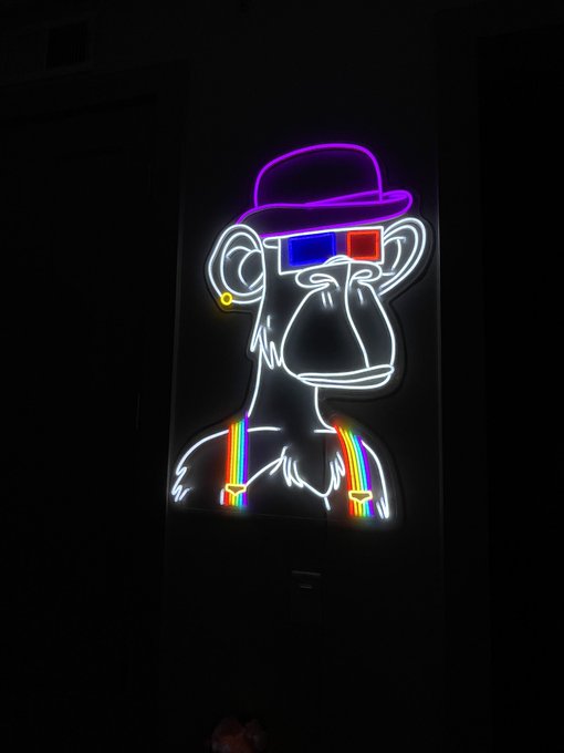A neon sign depicting a monkey wearing a purple hat and 3D glasses with rainbow suspenders, glowing against a dark background.