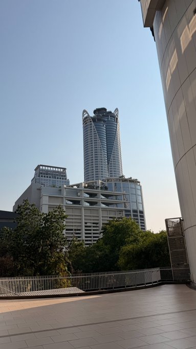 A tall modern skyscraper rises against a clear blue sky from what appears to be an urban plaza or rooftop terrace, with no visible text or signage. The grand architecture of the towering building pairs well with a "GM" greeting, symbolizing an optimistic start to the day against a sleek cityscape.