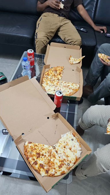 Two Domino's pizza boxes sit on a glass coffee table, each containing half a cheese pizza, accompanied by cans of Coca-Cola and a water bottle, as people relax on a couch and enjoy a casual weekend meal together.