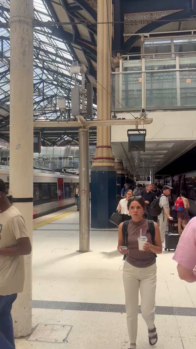 The image shows a busy train station with people walking around and waiting for their trains. A woman wearing a brown shirt can be seen holding a drink, possibly enjoying her time at the station or preparing to board her train. The photo also includes a watermark that reads "Gooood morning family! It's an even better one to IRL onboard Hey London, $retire is here! And not to forget on the best day of the week DCA Friday Let's have a great one you legends". This watermark suggests that the image may be related to a social media post or advertisement.