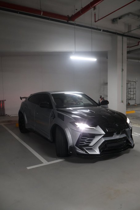 A gray modified Lamborghini Urus with aggressive body kit and illuminated headlights is parked in an underground parking garage.