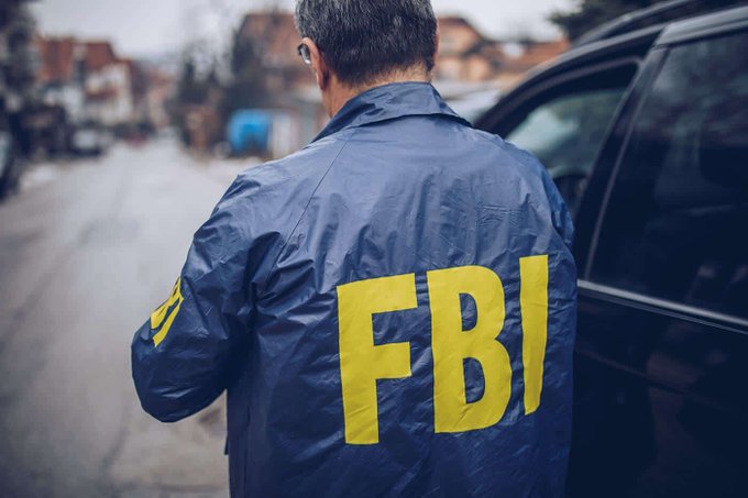 An FBI agent wearing a windbreaker with "FBI" in yellow lettering stands facing away from the camera next to a vehicle, illustrating the story of a federal agent who allegedly stole $1 million in cryptocurrency and consulted ChatGPT for advice on fleeing the country.