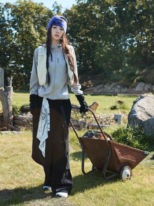 A person with long dark hair wearing a blue bandana, gray jacket, and black pants stands in a grassy outdoor area next to a wheelbarrow filled with items.