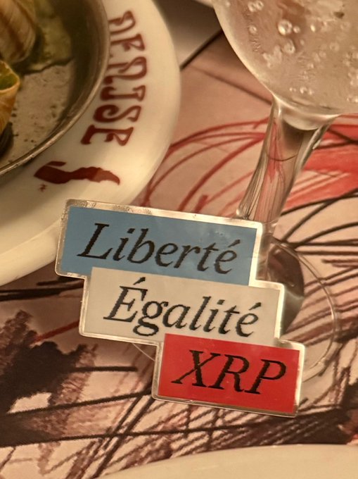 Three placards displaying "Liberté," "Égalité," and "XRP" rest on a table next to an overturned wine glass, cleverly replacing the French Revolutionary motto "Liberté, Égalité, Fraternité" with the cryptocurrency ticker to celebrate XRP as a third pillar.