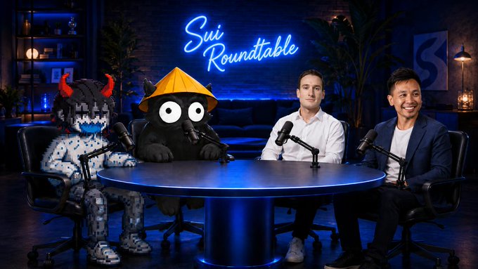 Two guests and two costumed mascots sit around a table on the set of "Sui Roundtable," a podcast-style show with a neon sign backdrop, as the hosts invite viewers to set a reminder.