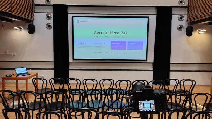 An empty conference room with rows of black chairs facing a projector screen displaying 'Zero to Hero 2.0 - LEARN CONNECT BUILD SHIP' with OCT 1-2, NEW MEXICO dates, and $20,000 pricing information.