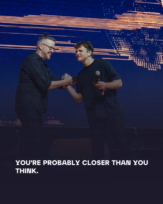 Two men in dark clothing shake hands on stage while holding microphones, with the caption "YOU'RE PROBABLY CLOSER THAN YOU THINK." displayed below.