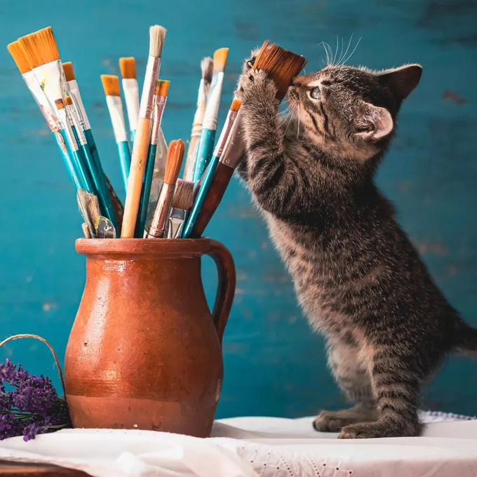 A curious tabby kitten stands on its hind legs, reaching up to play with a paintbrush from a terracotta pitcher filled with various artist's brushes, embodying the caption "Your artist" with its playful, creative spirit.