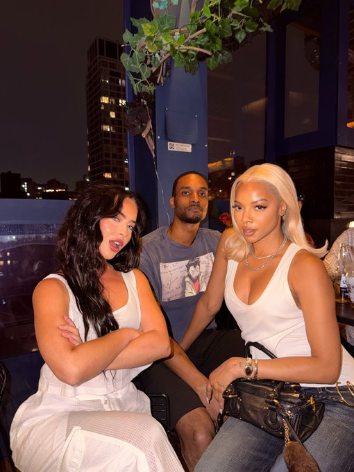 Three people pose together at what appears to be a rooftop venue at night, with a city skyline visible in the background; two women in white tops flank a man in a graphic t-shirt. The image shows a casual social gathering, suggesting "$ANSEM holders roll call" refers to a meetup or fan event for supporters of the Solana-based memecoin $ANSEM.