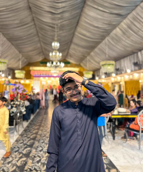 A young man wearing glasses and a dark blue traditional kurta stands in an elaborately decorated event hall with chandeliers, smiling while running his hand through his hair.