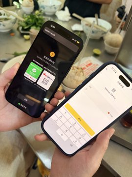 Two hands holding smartphones displaying what appears to be a mobile payment or messaging app interface, with a dining table and food visible in the background.