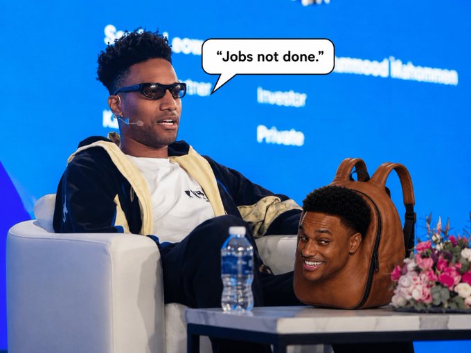 A panel discussion featuring a man in sunglasses with a speech bubble reading 'Jobs not done.', sitting next to a novelty backpack designed to look like a smiling man's head.