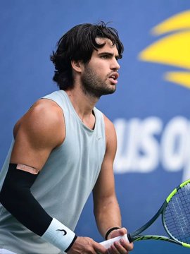 A focused tennis player with dark hair grips his racket on court, with "US O" visible in the background, suggesting a match at the US Open tournament. The image likely depicts Carlos Alcaraz, the player recently receiving an $8.4K bet on Polymarket to win the championship.