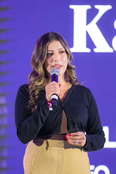 The image features a woman standing on a stage, holding a microphone and speaking into it. She appears to be addressing an audience or participating in a live event. The background of the image shows a blue wall with white text that reads "Chainlink hosting a full stage at ETHconf in NYC". This suggests that the woman is likely involved in an event related to technology or computer science, possibly at an academic conference or a tech-related gathering.