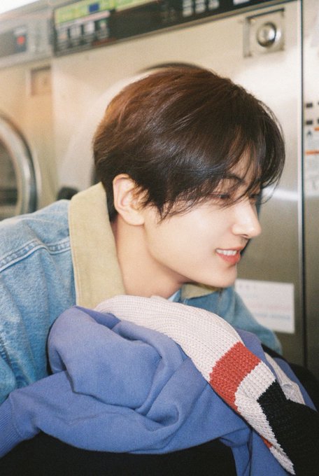 A young man with brown hair smiles in a side profile shot, wearing a denim jacket and holding a blue and red/white striped knit fabric, with laundromat machines visible in the background.