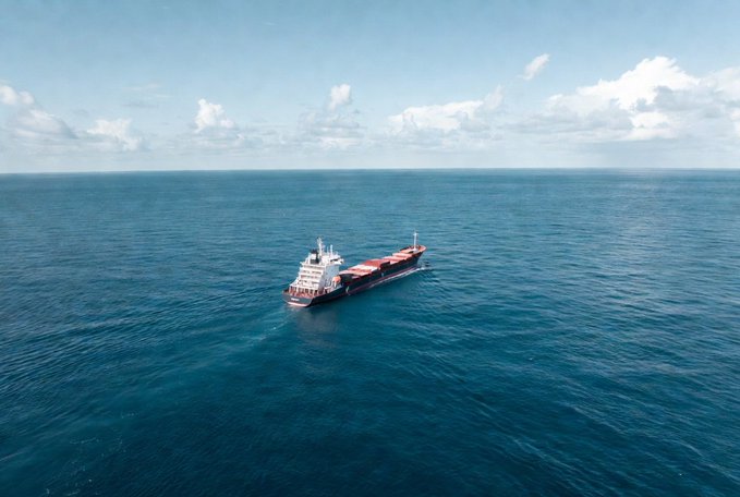 An aerial photograph of a large cargo ship sailing across a vast blue ocean under a partly cloudy sky.