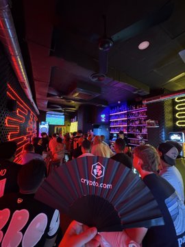 The image shows a large group of people gathered at a bar, possibly attending a Champions League event or watching a sports game. There are several individuals standing around, some holding drinks and others engaged in conversations. A person is holding up a black fan with the word "CRYTO" written on it, which could be related to the event taking place or simply an interesting decoration choice for the bar's atmosphere. The overall scene suggests that this gathering is centered around sports-related activities or events happening at the venue.