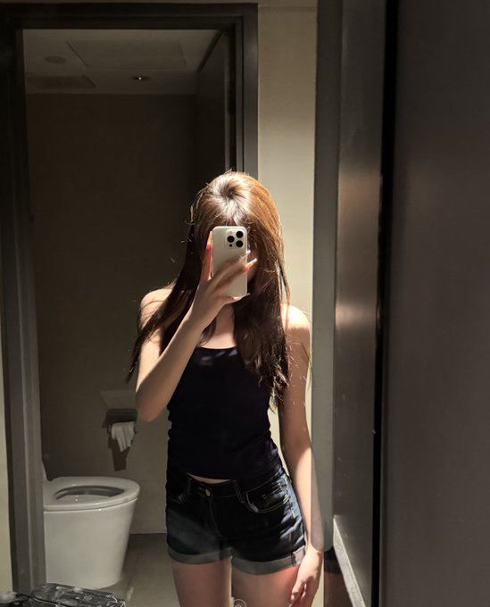 A mirror selfie of a young woman with long hair wearing a black tank top and dark denim shorts, holding a smartphone in front of her face inside a bathroom.