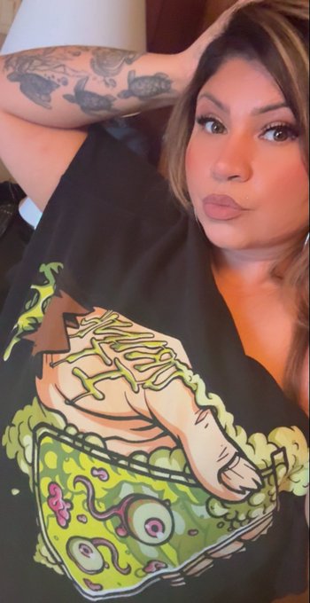 A selfie of a woman with tattoos wearing a black t-shirt featuring a colorful cartoon graphic of a hand emerging from a green oozing box labeled 'Venom'.