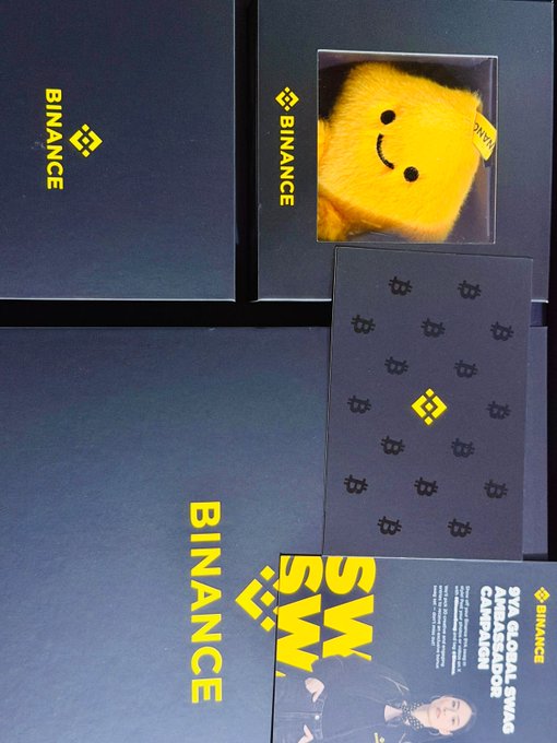 A 9-year anniversary Binance gift box is displayed, featuring branded dark packaging with the "BINANCE" logo, a plush smiley face toy visible in the top compartment, and an accompanying information card promoting the "BVA GLOBAL SWAG." The image showcases the premium quality merchandise received as a celebratory token from the Binance team.