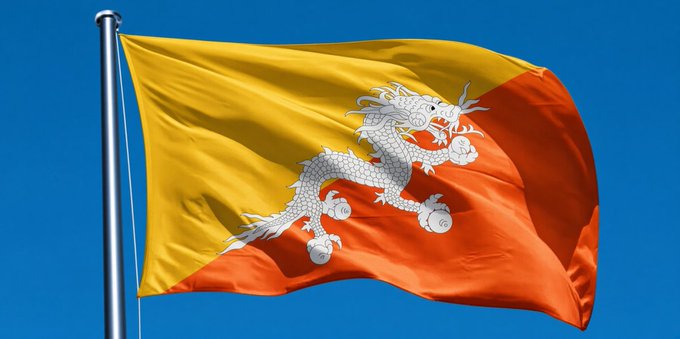 The image shows the national flag of Bhutan, featuring its distinctive yellow and orange halves divided diagonally with a white dragon in the center, flying against a blue sky. The flag relates to the post by visually identifying Bhutan, the country whose Prime Minister is being quoted about using Bitcoin to fund healthcare and public services.