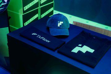 A promotional display featuring FXFlow branded merchandise, including a dark blue cap with the company's logo and folded apparel printed with the "FXFlow" name and matching logo, set against a vibrant green-lit backdrop. The setup reflects the brand's presence at the TOKEN2049 Singapore crypto event.