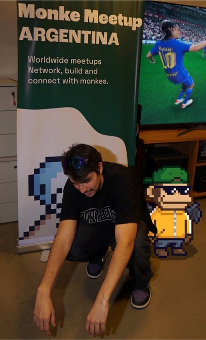 A person crouching next to a banner that reads 'Monke Meetup ARGENTINA - Worldwide meetups, Network, build and connect with monkeys,' with a pixelated Bored Ape character and a soccer player on a screen behind them.