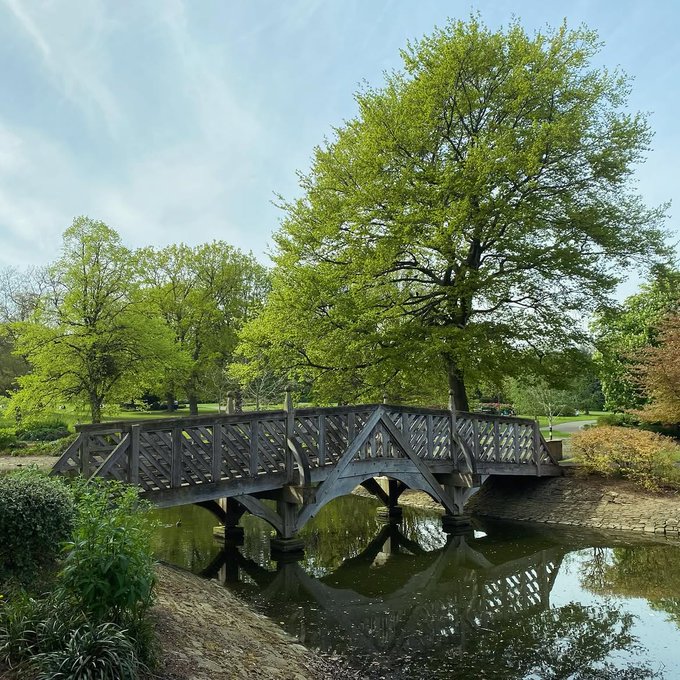 A wooden arched footbridge with lattice railings crosses a calm pond, surrounded by lush green trees and bushes under a partly cloudy blue sky.