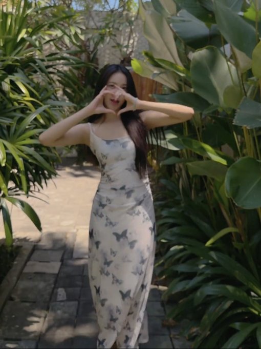 A young woman in a white butterfly-patterned dress makes a heart shape with her hands while standing on a path surrounded by tropical plants.