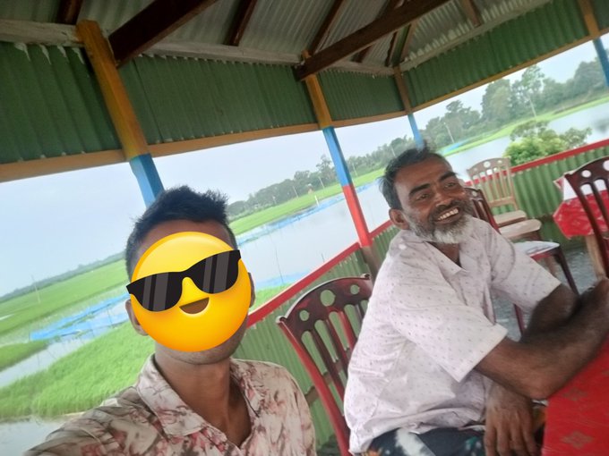 Two men sitting under a green-roofed shelter with a flooded field in the background, with one person's face obscured by a sunglasses emoji.