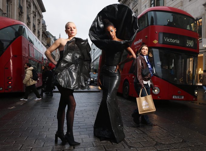 Two fashion models in avant-garde black outfits pose on a London street in front of a red double-decker bus marked 'Victoria 390', while pedestrians walk by carrying shopping bags.