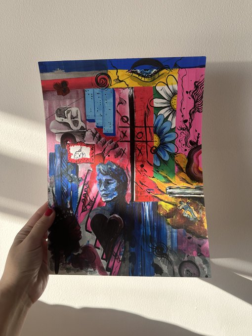 A hand holding up a colorful mixed-media painting featuring a melancholic blue character, bright flowers, a heart, and various abstract elements against a bright wall.