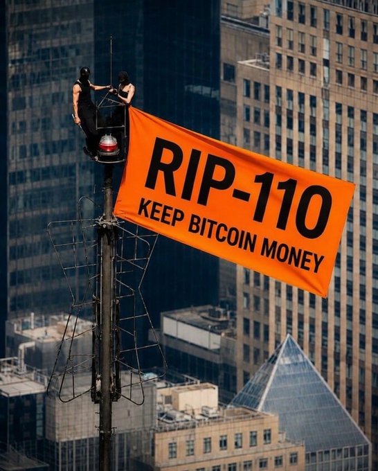 Activists in masks hang an orange banner reading 'RIP-110 KEEP BITCOIN MONEY' from a structure high above a city skyline.