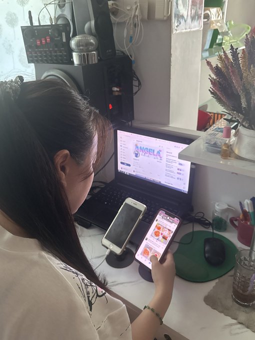 A young woman sits at a desk working on a laptop that displays the 'Angela' website, while she holds two smartphones in her hands.