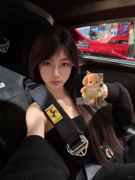 A young woman wearing glasses and a Ferrari seatbelt sits inside a red Ferrari, holding up a small plush toy in celebration of her grad school break and recent test drive of the new Ferrari F80.