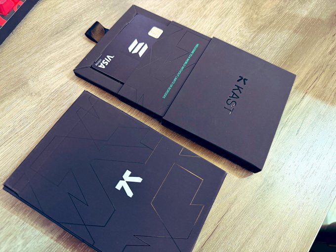 Two dark purple/black boxes from the brand 'KAST' on a wooden table, featuring geometric line designs and displaying a Visa card with teal text.