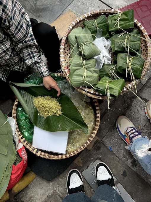 A street vendor selling leaf-wrapped sticky rice treats (zongzi) displayed in woven bamboo baskets, with a customer's hand reaching toward an unwrapped portion on a banana leaf.