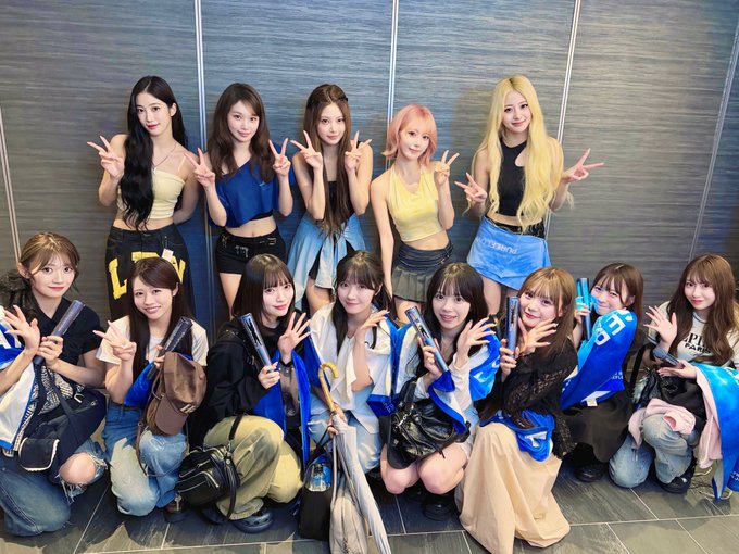 A group photo of around fifteen young women, mostly dressed in blue and white outfits, posing together and flashing peace signs indoors.
