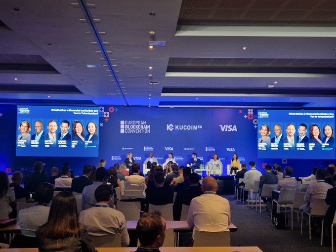 A conference room at the European Blockchain Convention filled with attendees facing a panel discussion stage, where the screen displays "What Inhibits a Financial Institution Key Prior to Tokenisation?" alongside KuCoin EU and Visa branding, contextualizing the live coverage of the institutional tokenization panel mentioned in the post.