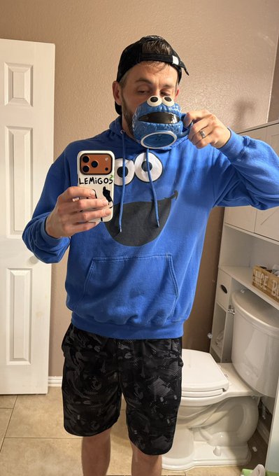 A man wearing a blue Cookie Monster hoodie, camo shorts, and a backwards cap takes a mirror selfie in a bathroom, with his phone case reading 'LEMIGOS'.