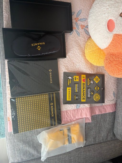 An unboxing photo showing Binance-branded merchandise including a sleep mask, notebooks, a sticker sheet, and a yellow item in plastic packaging laid out on a bed.