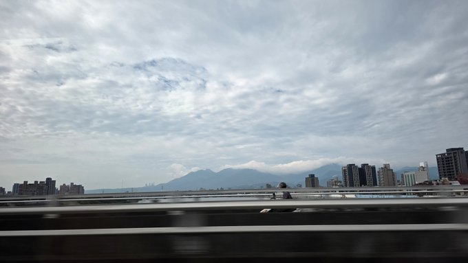 A cloudy, overcast sky blankets a distant cityscape and mountain range, viewed from a moving highway with blurred guardrails and two cyclists passing by. The gloomy, distant landscape mirrors the post's message—enduring storms of anger requires looking past the immediate turbulence toward a broader horizon.
