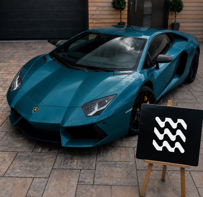 A teal/blue Lamborghini Aventador parked on a stone driveway in front of a wooden garage, displayed with a black sign featuring a white wavy line logo on a wooden easel.