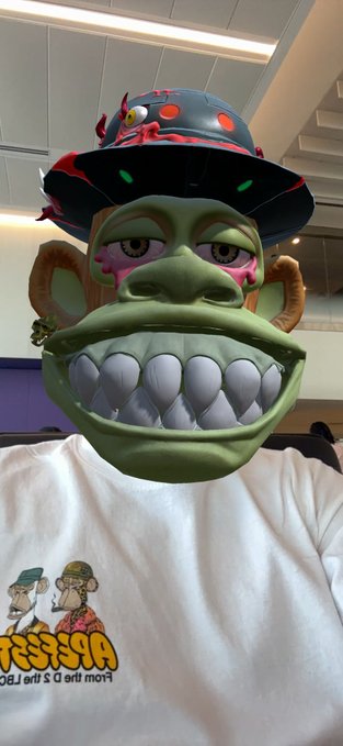 A person shows off a 3D Bored Ape Yacht Club-style mask/avatar with a green face, large toothy grin, and a red-accented hat, paired with a white t-shirt displaying the Bored Ape characters and mirrored text reading "BORED APE YACHT CLUB" — fitting the "GM" Sunday travel post en route to Chicago.