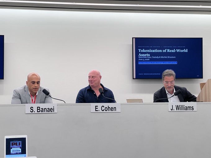 The image features a group of three men sitting at a table, with two men wearing suits and one man wearing a bald head. They are all seated behind microphones, suggesting that they might be participating in an event or panel discussion. The background includes a TV screen displaying information about tokenization law, which is likely related to the topic being discussed by the panelists.